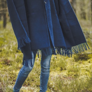 May include: A woman wearing a dark blue hooded poncho with fringe trim and blue jeans. She is standing in a forest setting.