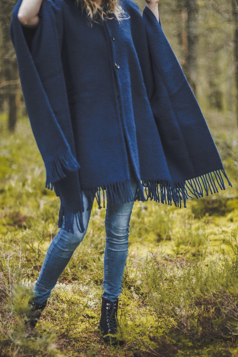 May include: A woman wearing a dark blue hooded poncho with fringe trim and blue jeans. She is standing in a forest setting.