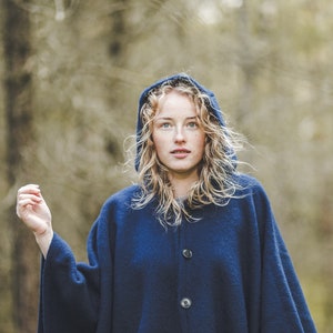 May include: A woman with long blonde hair wears a dark blue hooded cloak with a button closure. She is standing in a forest with a blurred background.