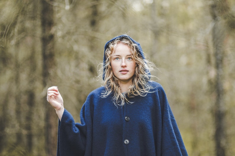 May include: A woman with long blonde hair wears a dark blue hooded cloak with a button closure. She is standing in a forest with a blurred background.