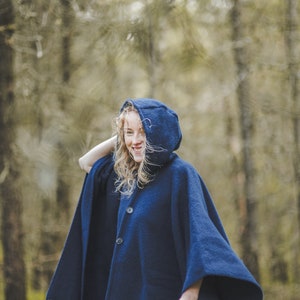 May include: A woman wearing a dark blue hooded poncho with fringe trim. She is standing in a forest and smiling.