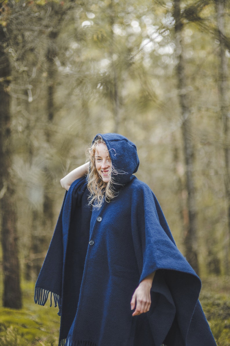 May include: A woman wearing a dark blue hooded poncho with fringe trim. She is standing in a forest and smiling.