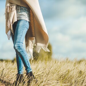 May include: A person wearing blue jeans and a beige fringed shawl walks through a field of tall grass.