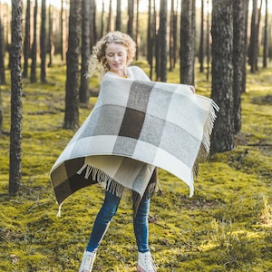 May include: A woman wearing blue jeans and white boots stands in a forest, holding a brown and white plaid blanket around her shoulders.