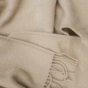 May include: A soft, beige throw blanket with a woven texture and fringe detail. The blanket is folded and draped over itself, showcasing the fabric's texture.