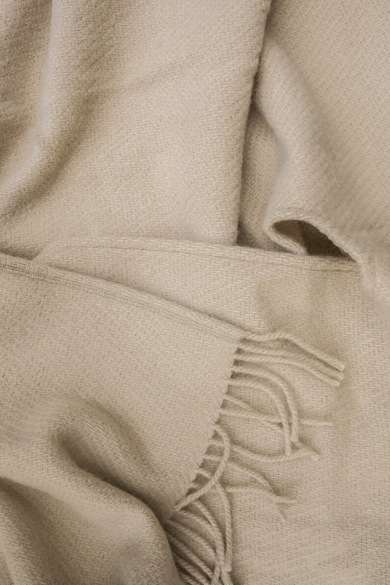 May include: A soft, beige throw blanket with a woven texture and fringe detail. The blanket is folded and draped over itself, showcasing the fabric's texture.