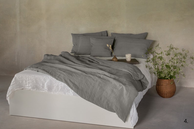 May include: A gray linen duvet cover and matching pillowcases are displayed on a white bed frame. The bed is against a textured gray wall. A small vase with dried flowers and a candle are on a wooden tray on the bed. A wicker basket with white flowers is on the floor next to the bed.