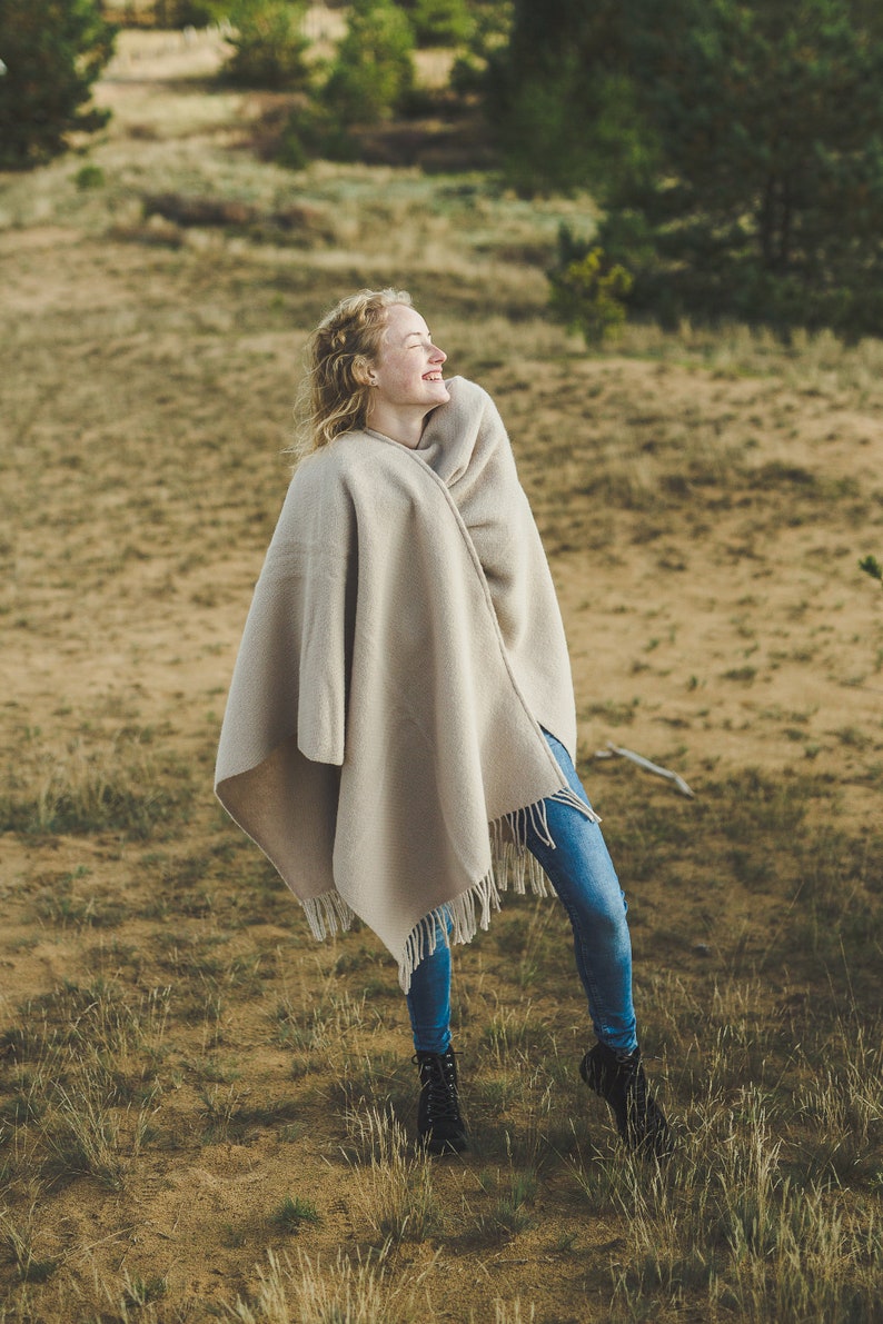 May include: A woman wearing blue jeans and a beige blanket wrapped around her shoulders smiles while standing in a field of tall grass.