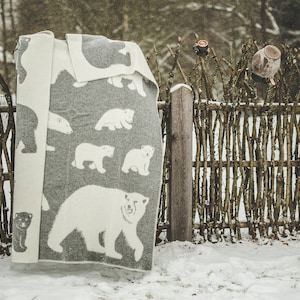 Könnte beinhalten: Eine graue und weiße gewebte Decke mit einem Muster aus Eisbären. Die Decke hängt an einem Holzzaun in einer verschneiten Winterlandschaft.
