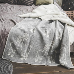Pode incluir: Uma manta de alpaca cinza com silhuetas de alpaca brancas. A manta está dobrada e drapeada sobre uma cama com uma manta de lã branca e cinza.