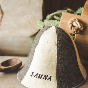 Puede incluir: Un gorro de sauna de fieltro blanco y marr&oacute;n con la palabra "SAUNA" bordada en la parte delantera.
