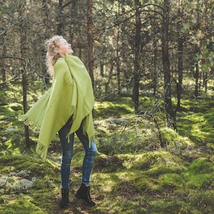 May include: A woman wearing a green shawl stands in a forest with green moss covering the ground. The woman is looking up and smiling.