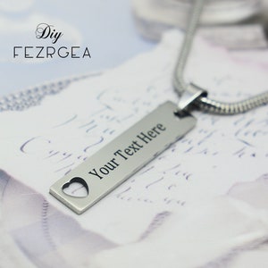 May include: A silver-colored pendant necklace with a rectangular tag featuring the text "Your Text Here" and a heart-shaped cutout. The necklace has a snake chain and the brand name "FEZRGEA" is visible.