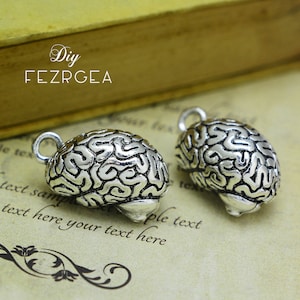 Può includere: Due ciondoli a forma di cervello color argento con dettagli intricati, perfetti per la creazione di gioielli o l'artigianato. Ogni ciondolo ha un anello per un facile fissaggio. Lo sfondo include testo e le parole "Diy FEZRGEA".
