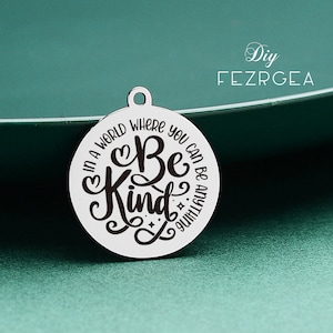 Puede incluir: Un amuleto de plata con las palabras "En un mundo donde puedes ser cualquier cosa, sé amable" en una fuente de letra cursiva negra.