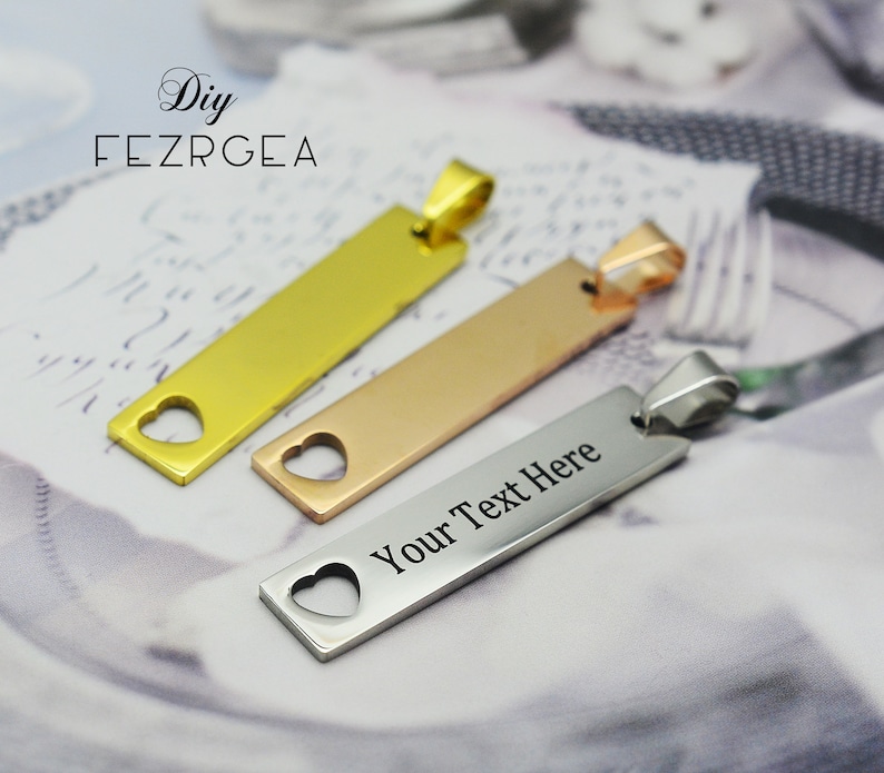 May include: Three rectangular pendants in gold, rose gold, and silver. Each pendant has a heart-shaped cutout and a loop for a chain. The silver pendant has the text "Your Text Here" engraved on it. The pendants are displayed on a patterned surface.