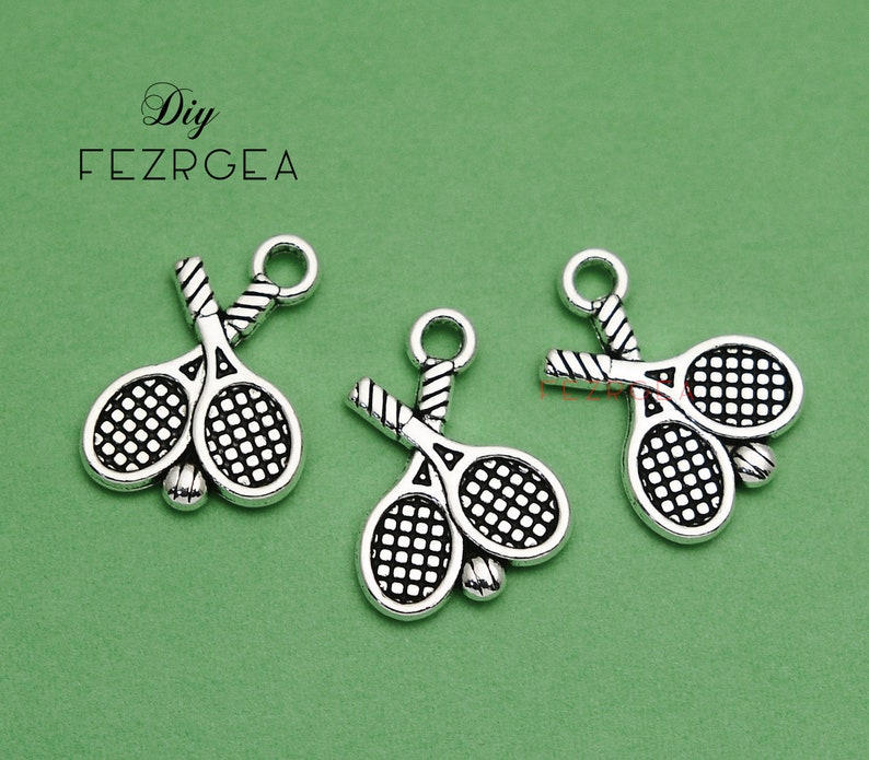 May include: Three silver-toned tennis racket charms with a black net design. The charms are hanging from a white background.