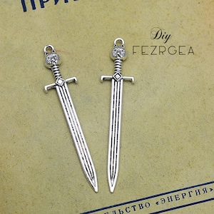 Può includere: Due ciondoli a forma di spada color argento con un design a testa di lupo sulla parte superiore. Le spade hanno un'elsa e una lama dettagliate, perfette per la creazione di gioielli o l'artigianato. Lo sfondo è carta vintage.