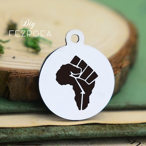 Puede incluir: Un charm de plata con un diseño en relieve negro de un puño levantado en el aire, con forma del continente africano. El charm es redondo y tiene un agujero en la parte superior para sujetarlo a una cadena o pulsera.