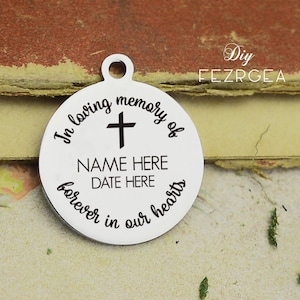 Op de afbeelding: Een ronde zilveren hanger met de tekst "In loving memory of" en een kruis boven de woorden "NAME HERE DATE HERE" en de tekst "forever in our hearts" eronder.