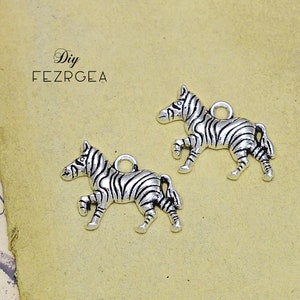 May include: Two silver-colored zebra charms with black stripes. Each charm has a loop at the top for attaching to jewelry or other crafts. The charms are on a tan background.