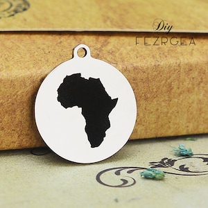 Puede incluir: Un charm de plata con una silueta negra del continente africano. El charm es redondo y tiene un agujero en la parte superior para sujetarlo a un collar o pulsera.