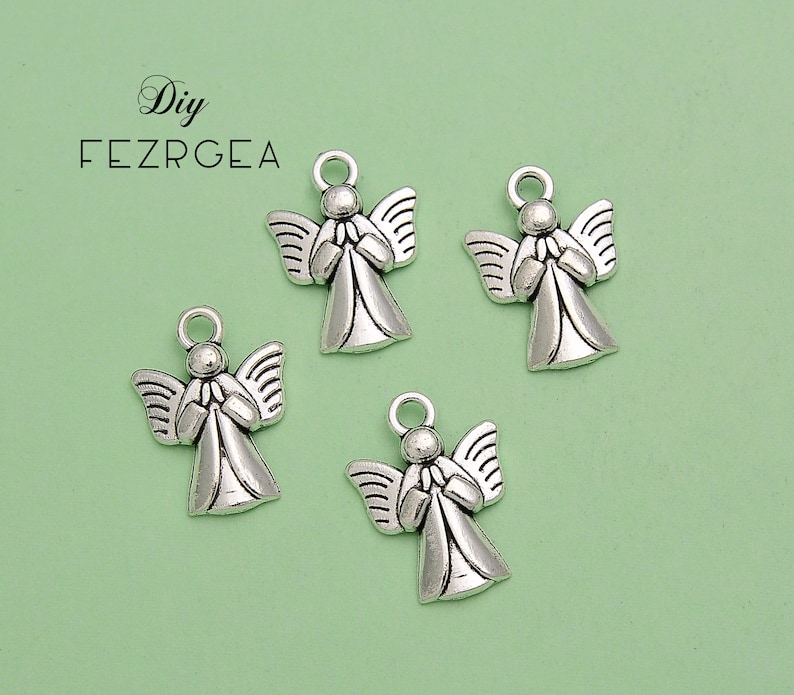 May include: Four silver-colored angel charms with detailed wings and clasped hands. Each charm has a small loop at the top for attaching to jewelry or other crafts. The background is a light green color.