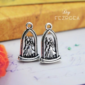 Puede incluir: Dos colgantes de plata con un par de novios de pie bajo un arco. Los colgantes están detallados con una novia y un novio en un escenario de ceremonia de boda.
