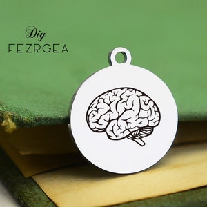 Puede incluir: Un amuleto de plata con un contorno negro de un cerebro humano. El amuleto es redondo y tiene un bucle en la parte superior para sujetarlo a un collar o pulsera.