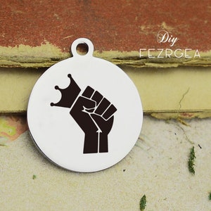 Puede incluir: Un amuleto de plata con una silueta negra de un puño levantado sosteniendo una corona. El amuleto es redondo y tiene un agujero en la parte superior para sujetarlo a un collar o pulsera.