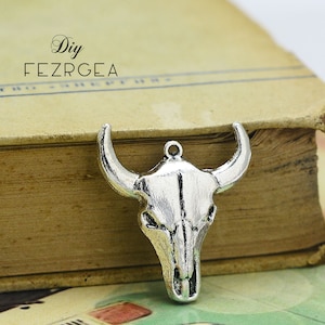 Peut inclure: Charme en métal argenté en forme de crâne de taureau avec un design détaillé. Le charme est suspendu à un livre avec le texte "DIY FEZRGEA" sur la couverture.