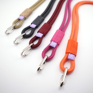 Op de afbeelding: Een verzameling kleurrijke lanyards in tinten beige, grijs, bordeauxrood, roze en oranje. Elke lanyard heeft een geweven stoffen gedeelte met een logo en een zilverkleurige metalen karabijnhaak.