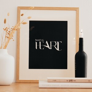 Könnte beinhalten: Ein gerahmter Schwarzweißdruck mit dem Text "head vs. HEART" in einer weißen serifenlosen Schrift. Der Rahmen ist aus hellem Holz und steht auf einem Holztisch mit einer weißen Vase und getrockneten Blumen, einer brennenden Kerze in einer schwarzen Flasche und einem Stapel Bücher.