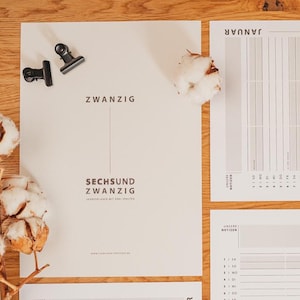 May include: A flat lay of a wooden surface with stationery and cotton stems. The stationery includes a white paper with German text and two black binder clips. There are also two other sheets of paper with calendar layouts. The cotton stems add a natural touch.