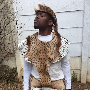 May include: A man wearing a traditional African garment with a leopard print pattern. The garment is made of fur and has a long, flowing tail. The man is also wearing a hat with a white feather.