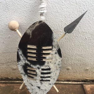 Puede incluir: Un escudo decorativo con un patrón moteado en blanco y negro, con una lanza y una bola de madera. El escudo tiene cortes horizontales y está montado sobre soportes de madera. Un elemento blanco y esponjoso está unido en la parte superior.