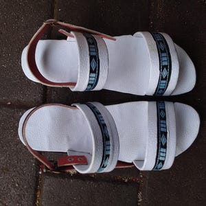 May include: Pair of white sandals with a textured footbed and straps. The straps feature a geometric pattern in blue and black. The ankle strap is red. The sandals are designed for casual wear.