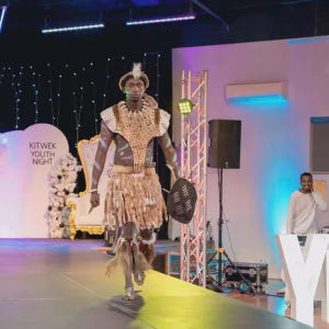 May include: A person in traditional African attire walks a runway. The outfit includes a beaded top, a skirt with fur-like trim, and a small shield. The event is labeled "KITWEK YOUTH NIGHT."