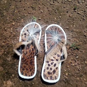 May include: A pair of sandals with a leopard print design and white trim. The sandals have a white strap and a white tassel on the top.