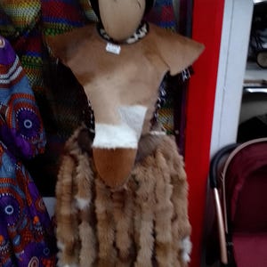 May include: A mannequin dressed in traditional African attire. The outfit features a brown and white hide top, a fur skirt, and a beaded necklace. A hat with a black brim and white top completes the ensemble. The outfit is displayed in a shop setting.