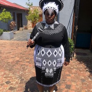 Può includere: Una donna che indossa un abito tradizionale sudafricano di perline. Indossa una gonna di perline nere e bianche, una blusa nera con un colletto di perline bianche e un grande cappello di perline bianche e nere con piume. Tiene un bastone di perline nella mano destra.