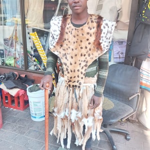 May include: A person wearing a traditional outfit with a leopard-print patterned cape and skirt, a feathered hat, and holding a wooden cane. The outfit is tan, brown, and white. The person is standing on a brick surface.
