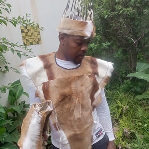 May include: A traditional outfit featuring a fur headdress with quill-like accents, a fur and hide chest piece, and a matching arm covering. The ensemble is primarily brown and white, with a white undershirt visible.