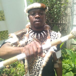 May include: A person wearing a traditional African outfit. The outfit includes a leopard-print top, a fur-trimmed collar with white teeth, and a white and brown hat. The person is holding a white and gold staff.