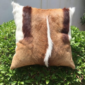 May include: A square pillow with a brown, white, and dark brown fur pattern. The pillow is sitting on a bed of green leaves.