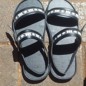 以下が含まれることがあります： 白い幾何学模様のストラップが付いた黒いサンダル2足。シンプルなデザインで耐久性のある素材で作られています。