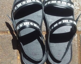 Könnte beinhalten: Ein Paar schwarze Sandalen mit weißen geometrischen Mustern auf den Riemen. Die Sandalen haben ein schlichtes Design und sind aus strapazierfähigem Material gefertigt.