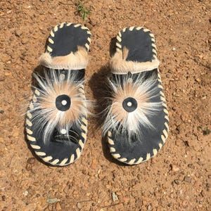 May include: A pair of black leather sandals with white trim and a fluffy white and brown pom-pom on each sandal.