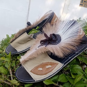 May include: A pair of handmade sandals with a unique design. The sandals feature a black sole, tan leather straps, and are adorned with a mix of brown and white fur. A brown leather bow adds a decorative touch. The sandals are displayed on a bed of green leaves.