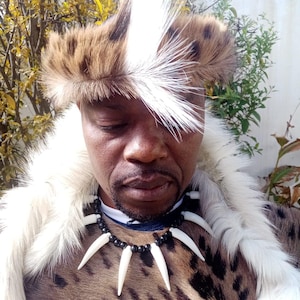 May include: A traditional African outfit featuring a spotted animal print garment, a fur hat with a white feather, and a necklace with white tusk-like pendants. The outfit includes a white fur collar.